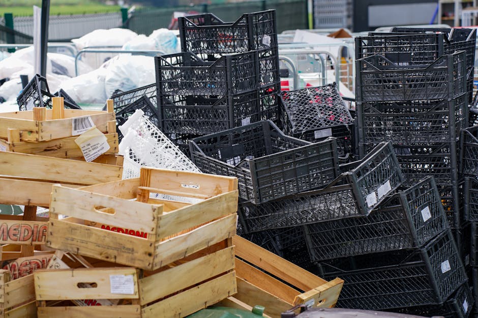 A large collection of black plastic crates and light wooden boxes stacked and piled outdoors, with some bins tilted or leaning against each other. The black crates have a lattice design, some containing smaller objects, while the wooden boxes appear empty, with visible slats and cutout handles. In the background, additional white plastic bags and other waste materials are partially visible, set against an urban environment with fencing and blurred structures. The scene is illuminated by natural daylight, creating a neutral, professional appearance suitable for describing a site involved in independent rubbish collection or on-site waste clearance. Rubbish Clearance Brixton could manage such waste, typical of bulk rubbish removal services after market days or during private disposal activities.