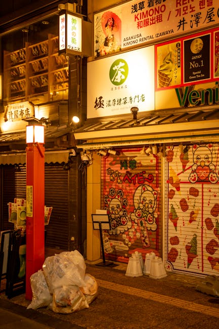 The image depicts the exterior of a commercial building at night, illuminated by warm ambient lighting. In the foreground, there are several plastic rubbish bags filled with waste, placed on the pavement near a red-painted vertical support beam. The background features a row of metal rolling shutters, with some partially closed, indicating commercial premises. The shutters are decorated with colorful, playful illustrations of cartoon characters, possibly pandas or similar animals, rendered in a bright red, orange, and pink palette, with some elements featuring speech bubbles. Above the shutters, there are illuminated signs in multiple languages, including Japanese and Korean, advertising various businesses such as a kimono rental shop and other establishments. The scene captures an urban setting with visible commercial activity, subtly reflecting the type of private waste disposal or on-site clearance services provided by Rubbish Clearance Brixton, in keeping with the rubbish removal theme of the page. The environment suggests a busy street area, possibly after business hours, with a focus on street-level waste awaiting collection or disposal.
