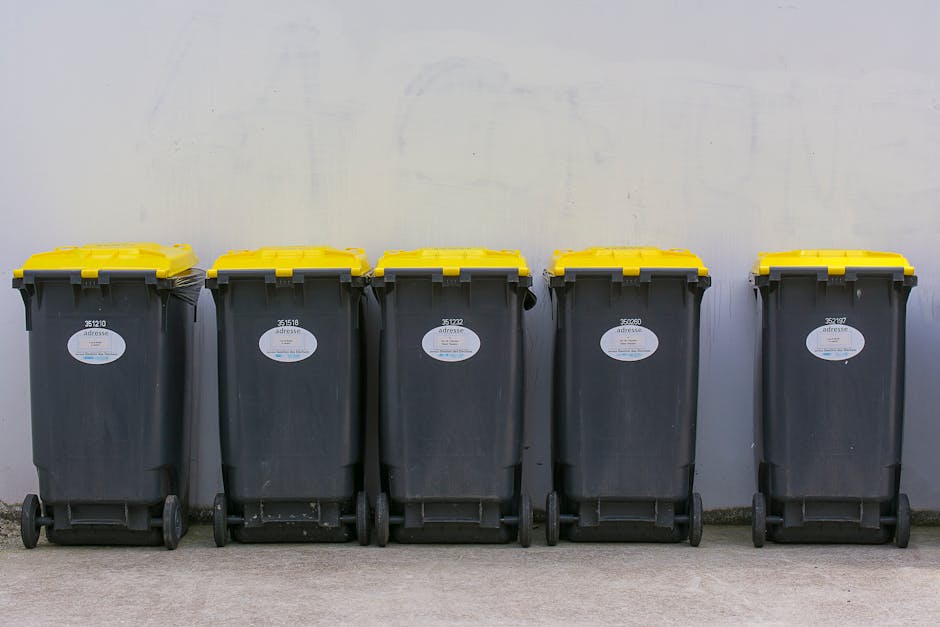 Five large wheeled rubbish bins with black plastic bodies and bright yellow lids are lined up against a plain, light-colored wall on a concrete surface. The bins are positioned side by side in a neat row, with each bin featuring a white label with black text and a small blue symbol. The environment appears to be either a dedicated rubbish collection area or a private waste disposal site, possibly associated with a commercial or residential property. The surface beneath the bins is smooth, and the overall scene is well-lit with natural or neutral lighting, emphasizing clean, functional waste management equipment. This setup may relate to independent rubbish clearance services provided by companies such as Rubbish Clearance Brixton for business or shop rubbish removal, illustrating organized private waste collection outside a building setting.