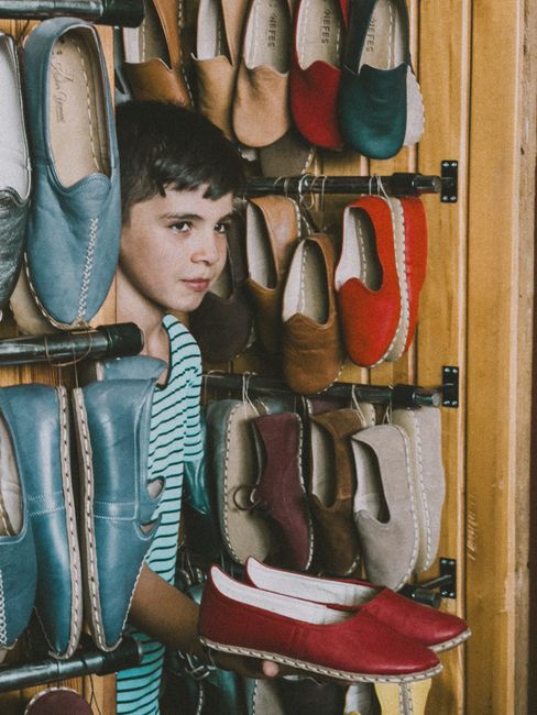 A young girl with short dark hair and a neutral expression is depicted in a shoe shop, standing among a display of various footwear. She is wearing a light blue and white striped top, and her face is partially framed by the array of shoes surrounding her. In front of her, she holds a pair of red slip-on shoes with a beige sole, featuring a smooth finish and a low, flat heel. Behind her, a wall-mounted rack displays numerous pairs of shoes, including flats, loafers, and slip-ons, arranged along horizontal metal rods. The shoes exhibit a variety of materials such as leather and synthetic fabrics, with finishes ranging from matte to slightly shiny, and in a range of colours including blue, red, beige, brown, and grey. Some shoes are stacked or leaning, creating a textured visual environment typical of a retail setting. The background consists of a wooden panel wall, adding warmth to the scene, and the lighting appears natural, softly illuminating the shoes and the girl's face. The image captures the context of footwear selection within a retail environment, aligned with private or independent shoe or rubbish disposal services, such as those provided by Rubbish Clearance Brixton in the context of waste management and rubbish removal services.