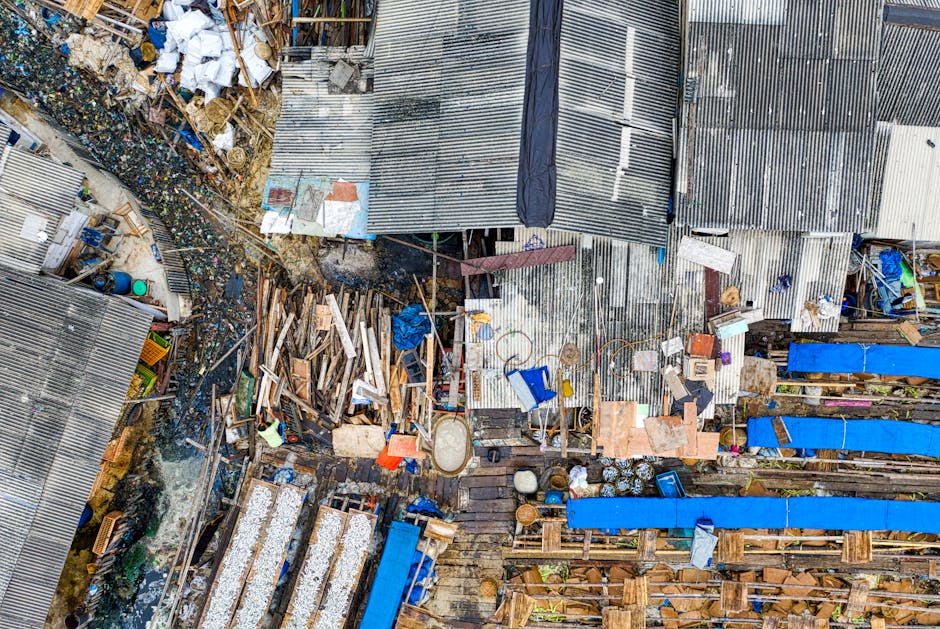 An aerial view of a densely cluttered outdoor area showing various piles of waste and construction debris scattered across uneven ground. The scene includes stacks of wooden planks, some broken or splintered, alongside corrugated metal sheets and roofing panels, mostly silver or grey with some blue tarpaulins draped over parts of the debris, indicating makeshift coverings or storage. The debris appears haphazardly arranged, with loose planks, plastic containers, and other waste materials visible, some resting on stacked pallets or amidst ongoing clearing efforts. To the left, a small section of a concrete pathway or driveway is partially visible, edged by the waste piles, while the background reveals more collapsed structures or sheds also constructed from corrugated metals. Bright natural lighting highlights textures of weathered wood, rusted metals, and scattered plastic waste, reflecting a scene related to independent waste handling or rubbish removal activities that could be carried out by Rubbish Clearance Brixton. The area's cluttered state conveys an ongoing process of rubbish collection and site clearance in an informal setting, suitable for private disposal or alternative waste management services.