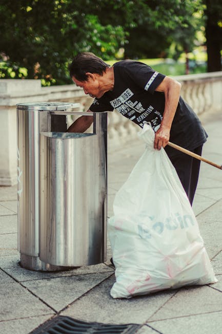 A woman dressed in a black T-shirt with white text and black pants is standing outdoors on a paved area near a curved stone railing. She is holding a large white plastic bag filled with rubbish in her left hand and is leaning forward, placing waste into a modern, stainless steel outdoor rubbish bin with a sleek, curved design and multiple compartments. The environment features lush green trees and grass in the background, suggesting a park or residential area. The scene is well-lit with natural daylight, creating a clear and detailed visual of the waste disposal activity, which reflects the importance of private rubbish collection services such as those provided by Rubbish Clearance Brixton for on-site waste management and alternative disposal options.