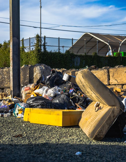 An aerial view of a densely cluttered outdoor area showing various piles of waste and construction debris scattered across uneven ground. The scene includes stacks of wooden planks, some broken or splintered, alongside corrugated metal sheets and roofing panels, mostly silver or grey with some blue tarpaulins draped over parts of the debris, indicating makeshift coverings or storage. The debris appears haphazardly arranged, with loose planks, plastic containers, and other waste materials visible, some resting on stacked pallets or amidst ongoing clearing efforts. To the left, a small section of a concrete pathway or driveway is partially visible, edged by the waste piles, while the background reveals more collapsed structures or sheds also constructed from corrugated metals. Bright natural lighting highlights textures of weathered wood, rusted metals, and scattered plastic waste, reflecting a scene related to independent waste handling or rubbish removal activities that could be carried out by Rubbish Clearance Brixton. The area's cluttered state conveys an ongoing process of rubbish collection and site clearance in an informal setting, suitable for private disposal or alternative waste management services.